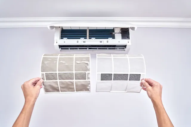 Hands holding a dirty air conditioner filter next to a clean one in front of a wall-mounted AC unit.