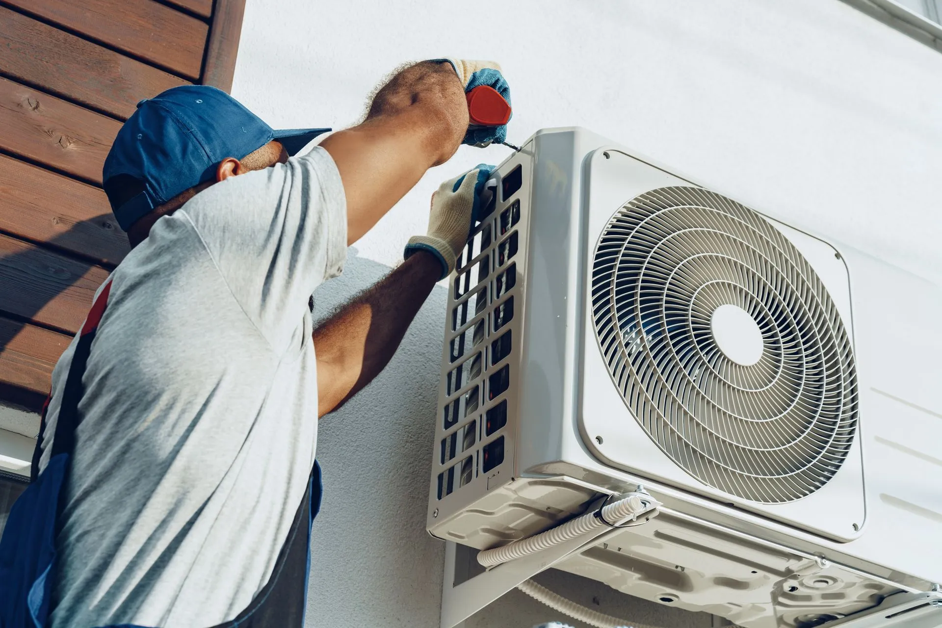 A worker in a blue cap and work clothes uses a screwdriver to service an outdoor air conditioning unit on a white wall.
