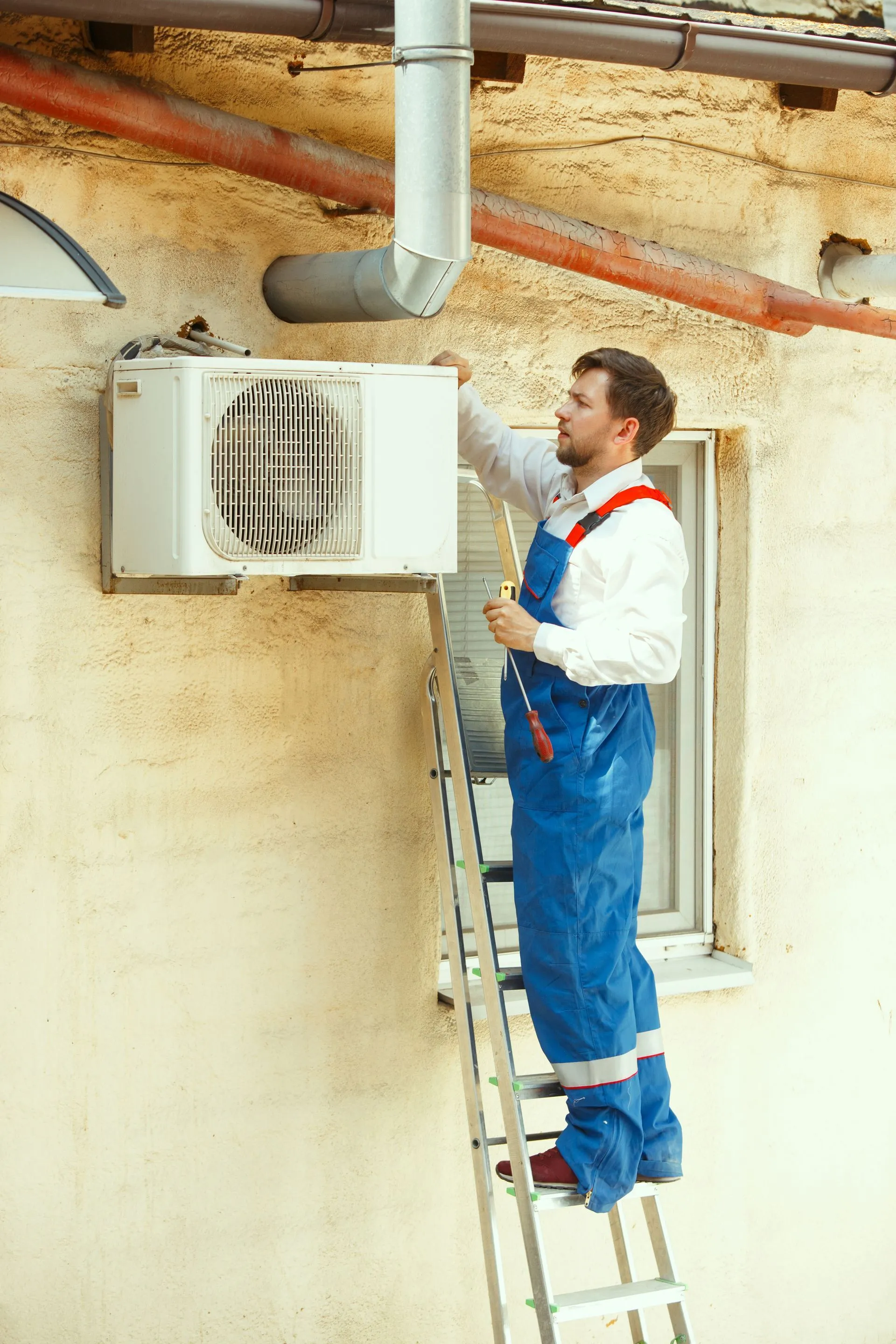A technician in blue coveralls stands on a ladder, installing an air conditioning unit on an exterior wall.