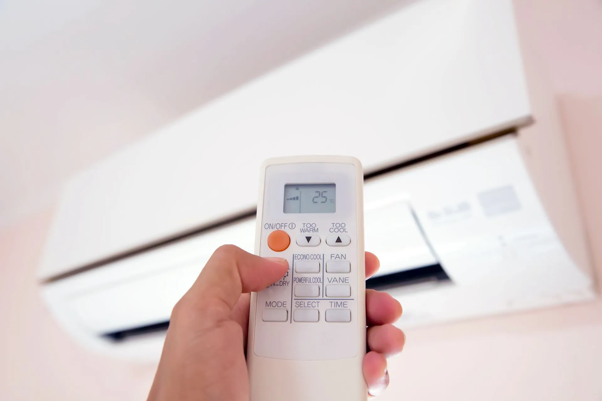 A hand holds a white air conditioner remote control pointing toward a wall-mounted indoor AC unit.